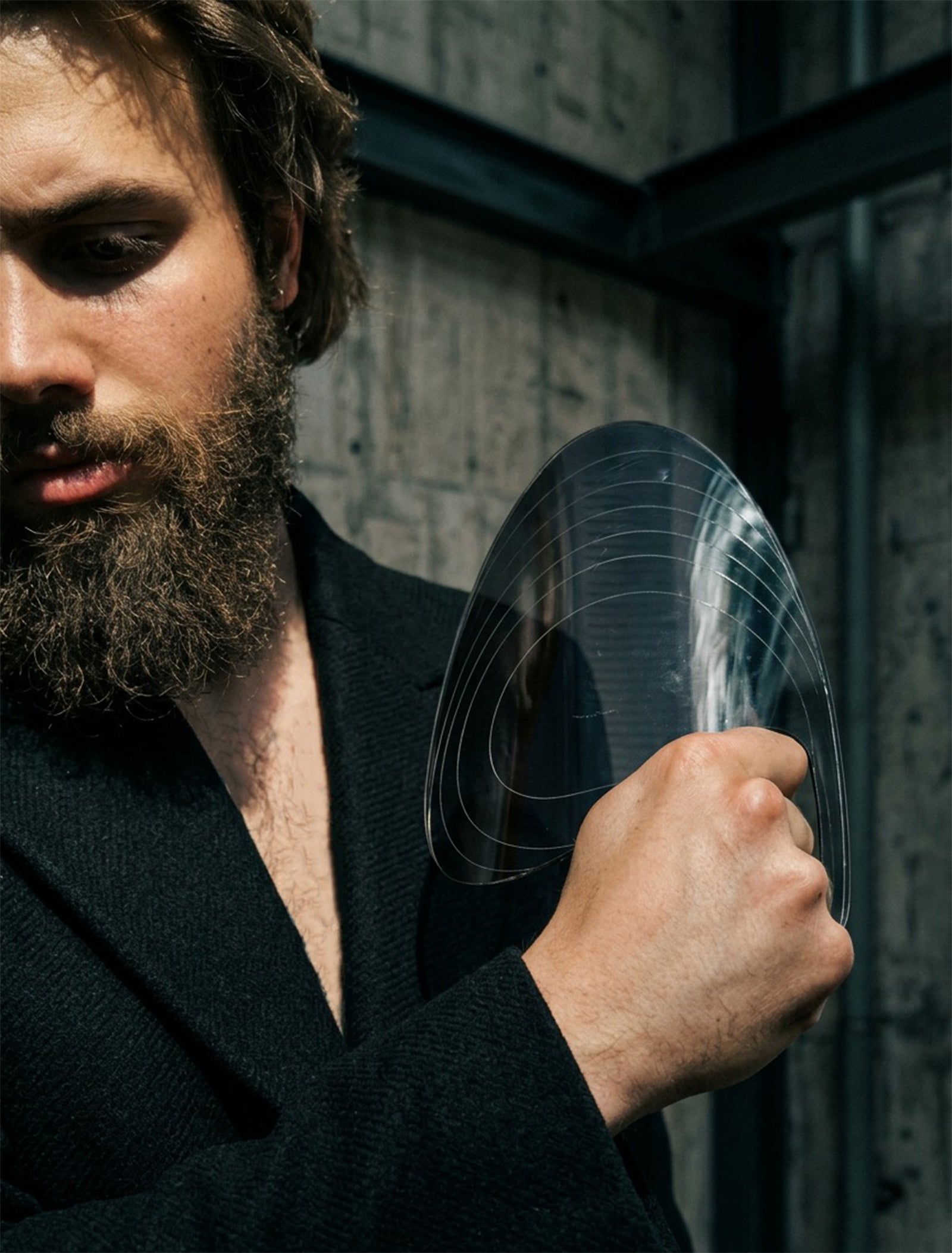 A bearded man in a dark wool coat holds the PRIOSA Prima clear acrylic accessory near his face, photographed in a brutalist concrete interior
