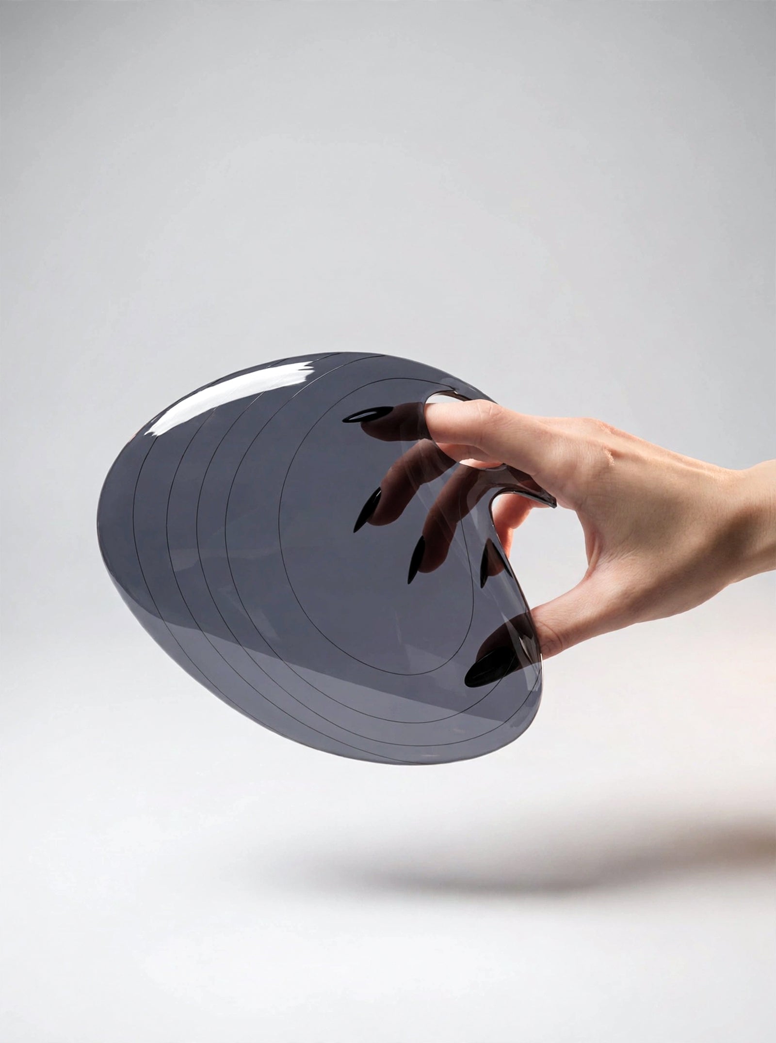 A feminine hand with glossy black almond-shaped nails holds the PRIOSA Prima smoke grey translucent acrylic design object, glossy reflective surface catching studio light, against a neutral grey studio backdrop